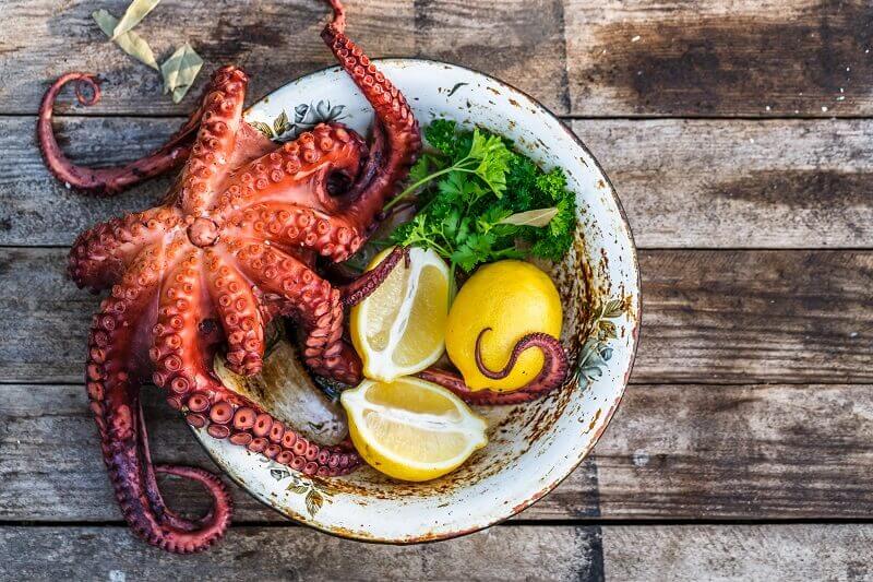 A whole grilled octopus sitting in a chipped white bowl with sprigs of parsley and lemon wedges. It is sitting on a wooden table outside.