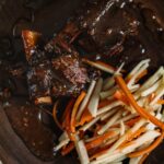 Braised beef short ribs on a wooden plate next to an apple and carrot slaw salad.