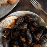 A plate of mussels in a garlicky red wine sauce with tomatoes and pancetta. A piece of bread is also on the plate.