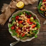 A big bowl and plate on a wooden table full of salad covered in roasted vegetables and dressing.