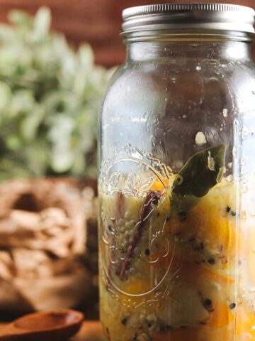 A large glass jar half full of muddled, smushed sliced meyer lemons and other ingredients like herbs and spices. The jar is getting ready to be fermented and preserved. It is on a wooden table and small white flowers can be seen behind.