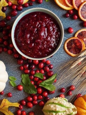 a bowl of freshly made cranberry sauce on a blue background surrounded by fall decor like pumpkins, wheat, and fresh cranberries
