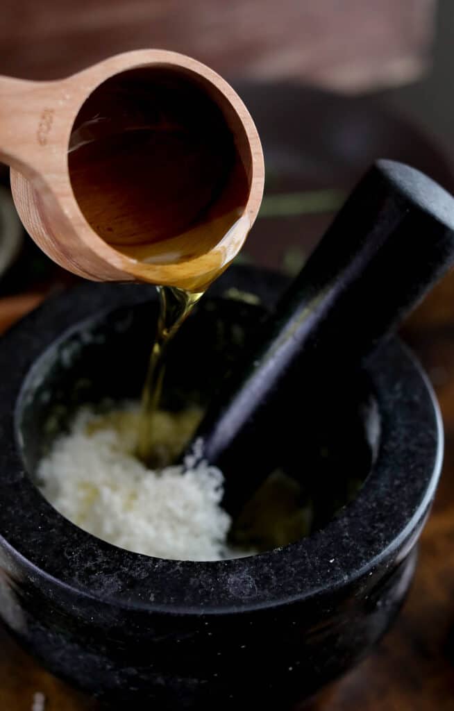 Olive oil being poured from a wooden measuring cup into a mortar full of pesto and cheese.