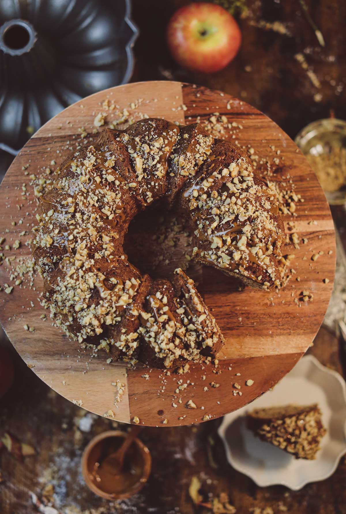 The top view of a fresh apple cake covered in dulce de leche caramel and crushed nuts. It is sitting on a wooden cake stand with a slice missing.