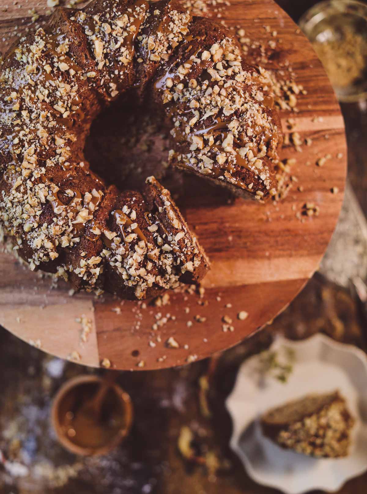 The top view of a fresh apple cake covered in dulce de leche caramel and crushed nuts. It is sitting on a wooden cake stand with a slice missing.