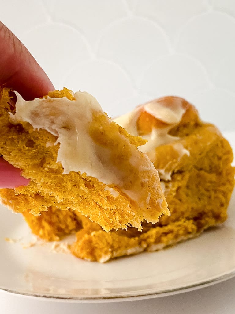 a close up shot of an individual pumpkin cinnamon roll topped with cream cheese frosting being pulled apart by a hand