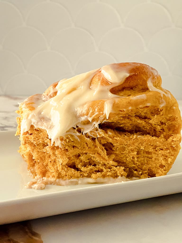 a close up shot of an individual pumpkin cinnamon roll topped with cream cheese frosting on a white plate