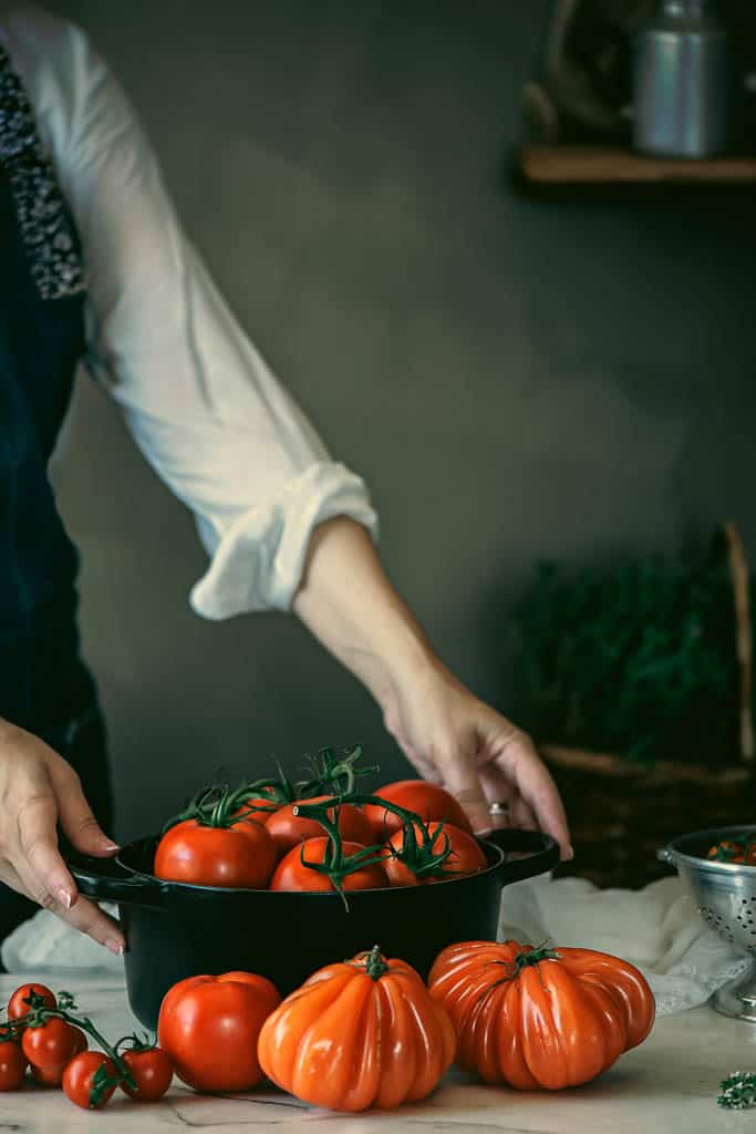 womans hands and arm in a white shirt holding a pot of red tomatoes on a marble kitchen countertop