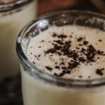 Two mugs full of warm vanilla custard milk bedtime drink. They are on a dark wooden tray with grated chocolate served on top.