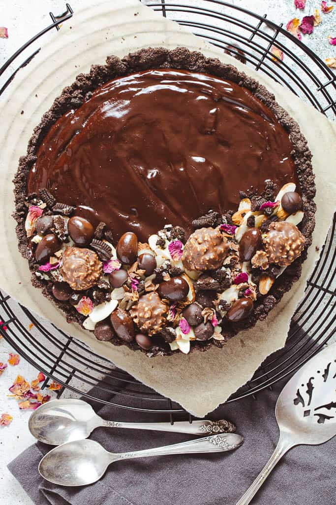 A chocolate tart decorated with ferrero rocher, chocolate covered almonds, broken cookie pieces, and dried rose petals. It sits on a wire rack.