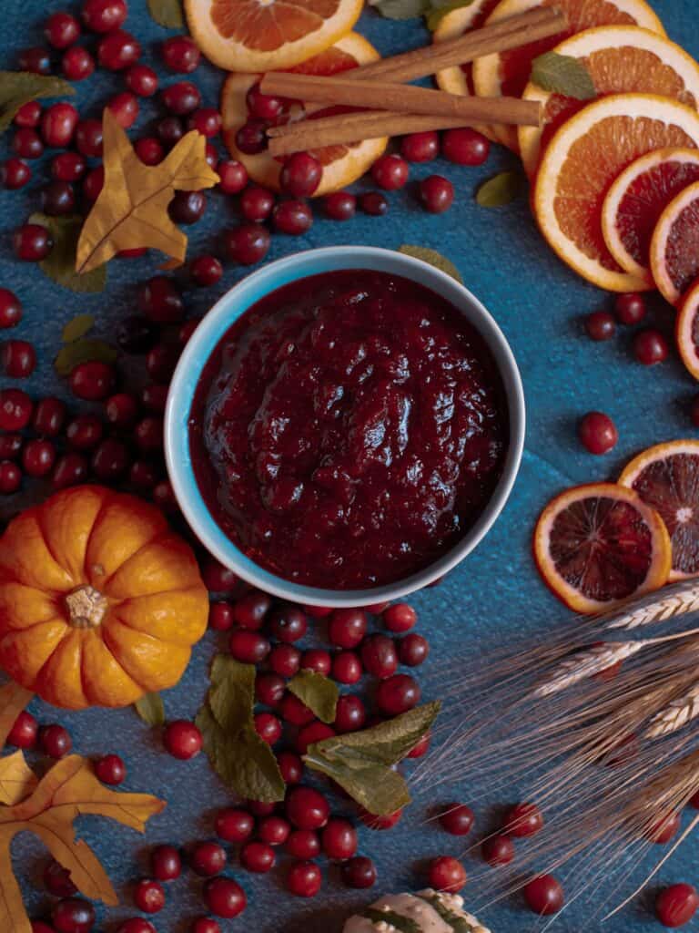 a bowl of smoked cranberry sauce on a dark blue background surrounded by fall decorations like pumpkins, wheat, fresh orange slices, and fresh cranberries