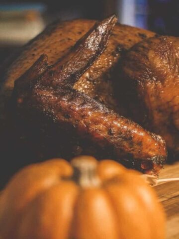 a browned and perfectly smoked whole turkey on a wooden board. In front is an out of focus pumpkin.