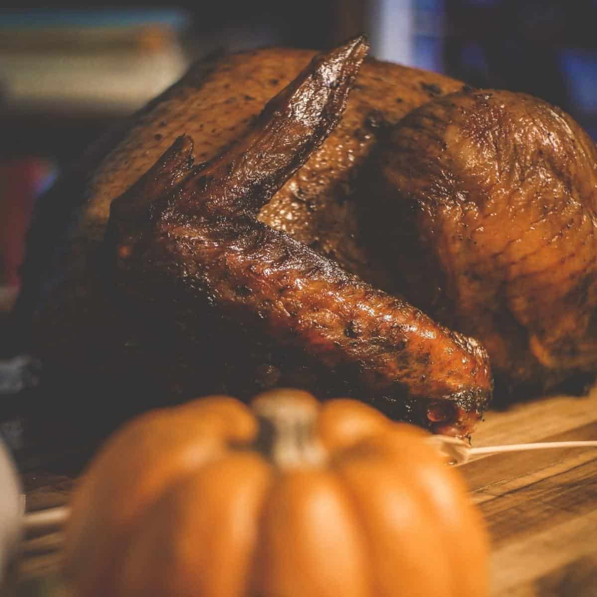a browned and perfectly smoked whole turkey on a wooden board. In front is an out of focus pumpkin.