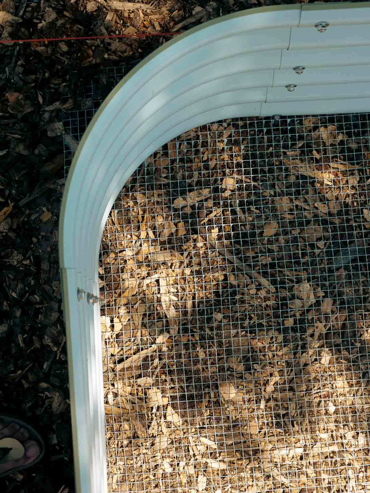 Close up of a green metal raised bed. It is empty but underneath it is hardware cloth and it is sitting on top of a thick bed of wood chips.