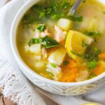 A bowl of Ukha fish soup with chunks of salmon, bream, and trout with vegetables like carrots and potatoes in a white bowl on a wood table.