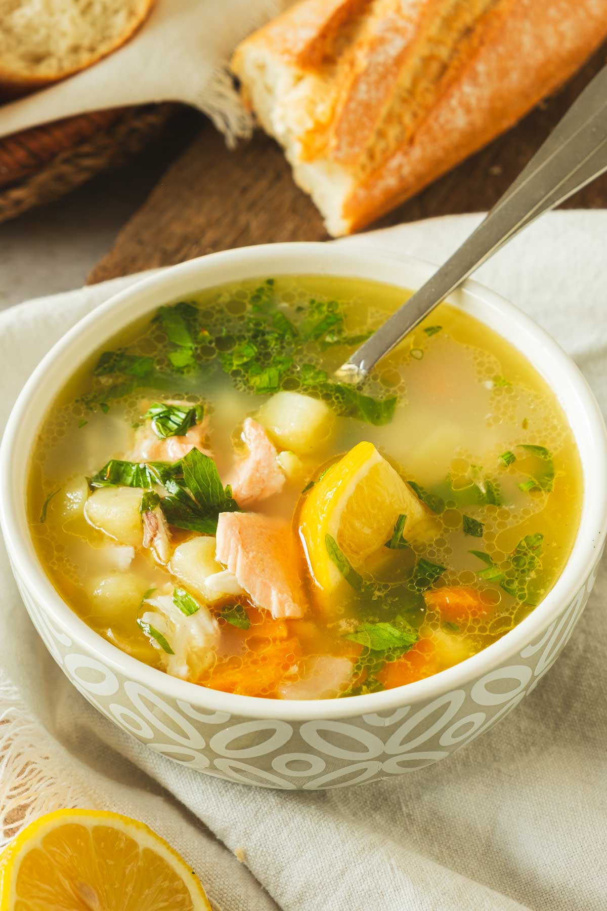 A bowl of Ukha fish soup with chunks of salmon, bream, and trout with vegetables like carrots and potatoes in a white bowl on a wood table.