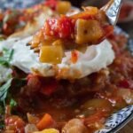 A blue and white plate heaped with sataraš, a medley of stewed or braised tomatoes, onions, and peppers in olive oil. It is served next to rice and with a fried egg on top. A fork is holding a piece of egg with the vegetables.