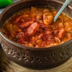 A large bowl full of Bosnian bean soup grah with a spoon in it. The soup is very thick and full of beans and other vegetables.