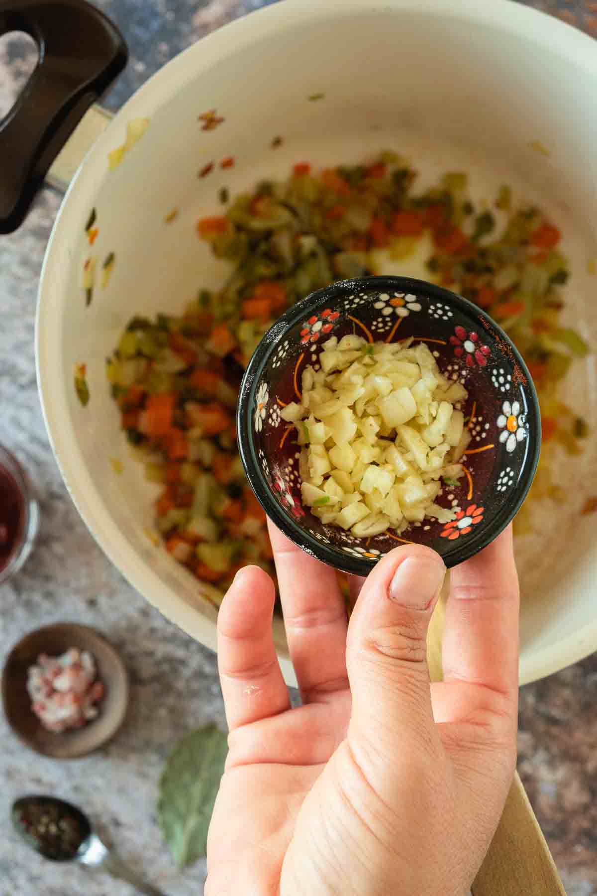 Minced garlic in small bowl being added to large pot full of diced, hot vegetables.