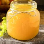 A large jar full of chicken feet bone broth. It is sitting on a blue linen napkin on a dark wooden table. The stock is golden yellow and very gelatinous.