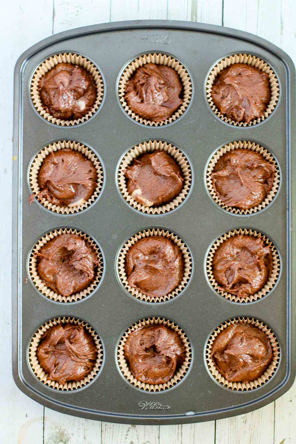 A 12-cupcake baking mold lined with cupcake wrappers and filled with raw chocolate peanut butter buckeye batter ready for baking.