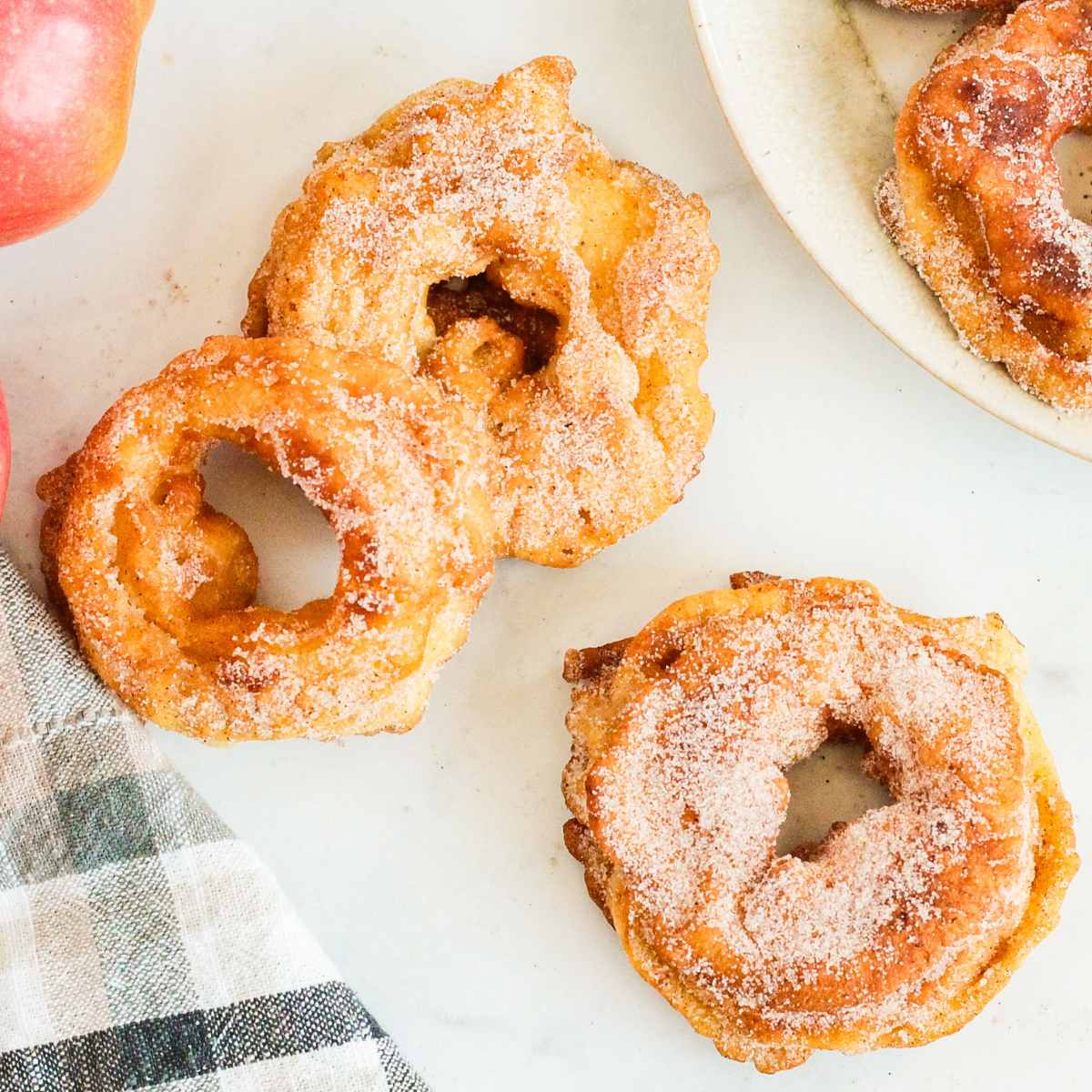 3 apple fritter rings on a white marble countertop next to a plate full of more apple rings. A plaid tea towel and fresh, red apples are next to them.