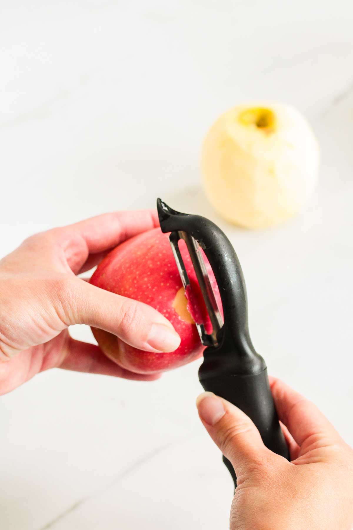 A red apple being peeled with a black vegetable peeler.
