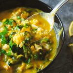 A black bowl full of golden glow soup. The soup is bright orange from the sweet potatoes and turmeric and very creamy. It is full of cooked rice, shredded chicken, and kale.