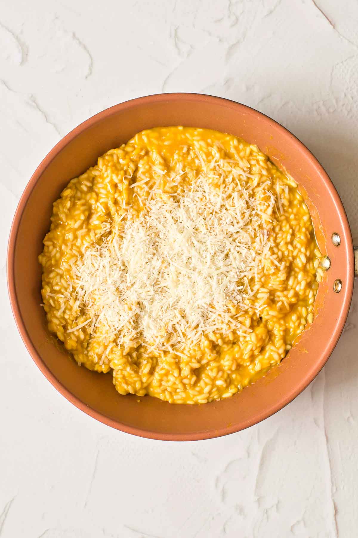 A big shallow bowl full of light-orange-colored pumpkin risotto. It is topped with a generous amount of Parmigiano Reggiano cheese.