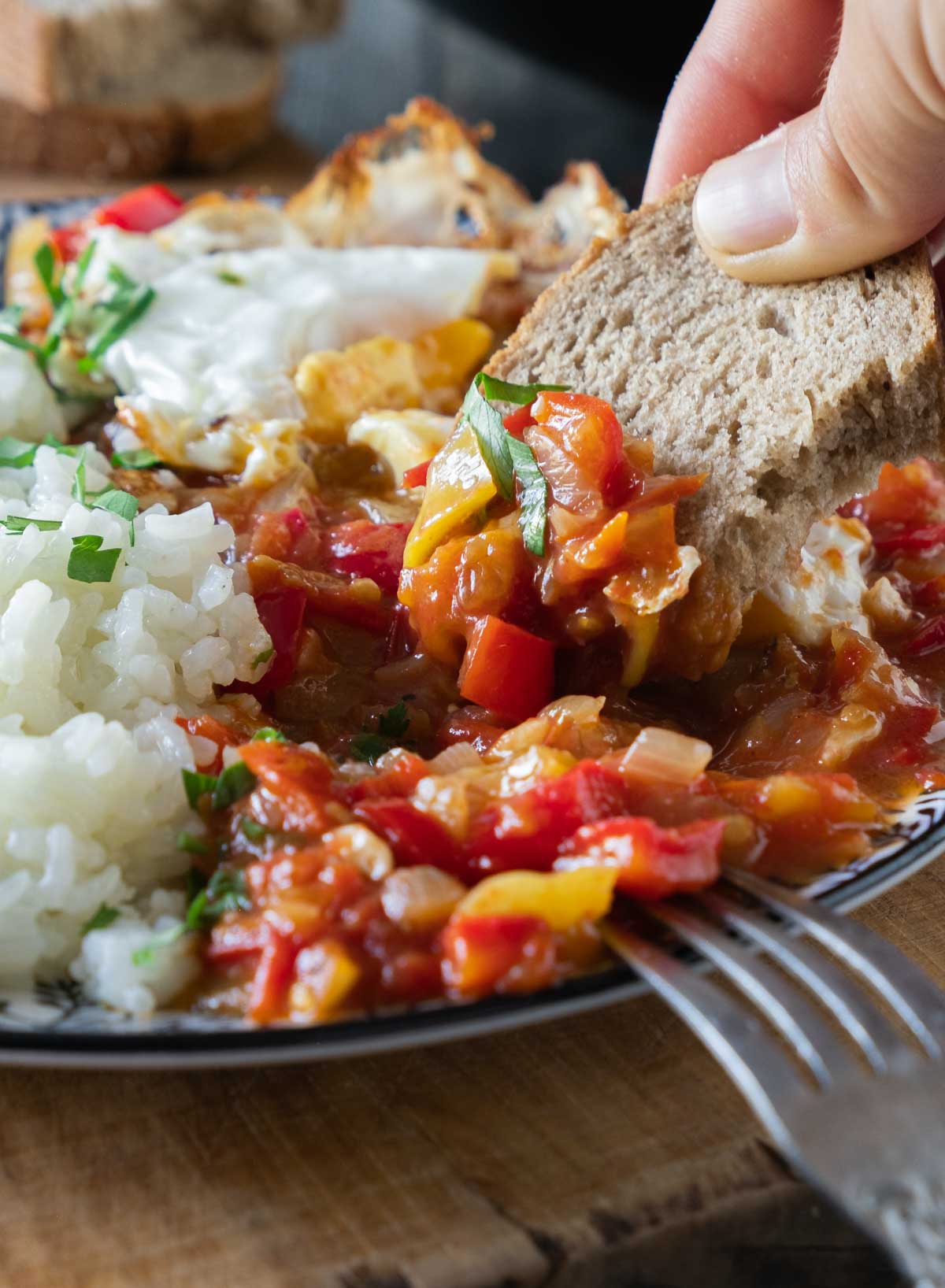 A blue and white plate heaped with sataraš, a medley of stewed or braised tomatoes, onions, and peppers in olive oil. It is served next to rice and with a fried egg on top. A piece of brown sourdough bread is being dipped into the sataras.