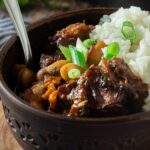 A large bowl full of Jamaican oxtail stew with butterbeans. Rice is served on the side and green onions are used as a garnish.