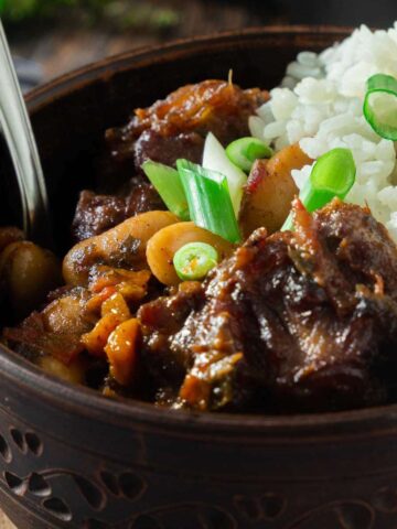 A large bowl full of Jamaican oxtail stew with butterbeans. Rice is served on the side and green onions are used as a garnish.