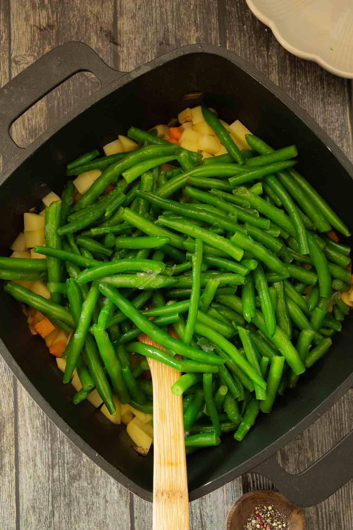 Long, fresh green beans being added into pot full of vegetables and water or stock.