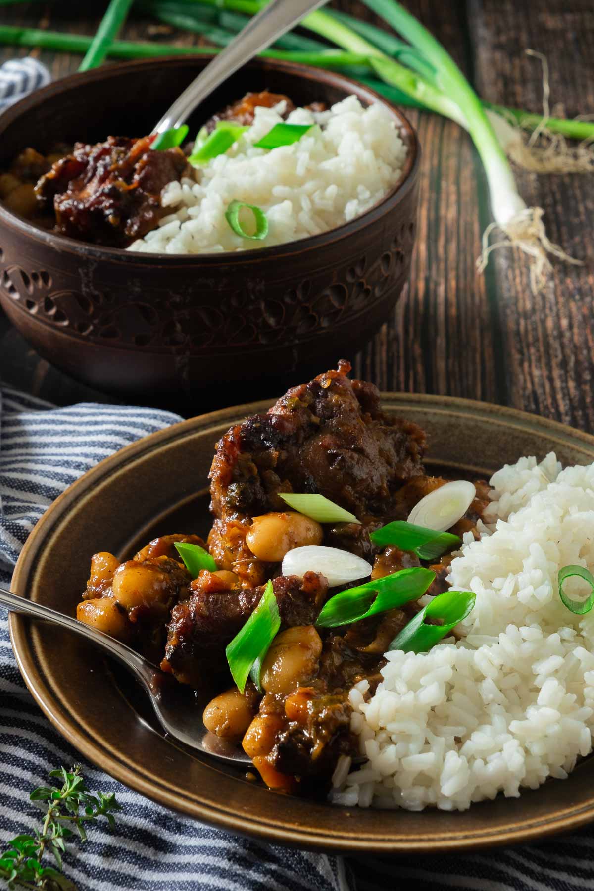 A large bowl full of Jamaican oxtail stew with butterbeans. Rice is served on the side and green onions are used as a garnish.