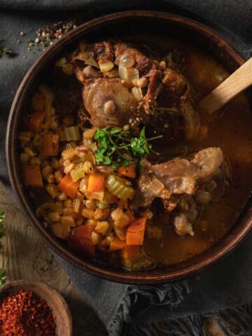 A clay bowl full of oxtail soup. There are whole chunks of oxtail in a rich broth with barley and diced vegetables.