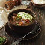 A clay bowl full of chunky red cabbage and winter root vegetable soup. There is a big dollop of sour cream on top.