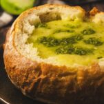 Roasted green tomato soup served in a sourdough bread bowl on a plate on a wooden table. There is pesto drizzled onto the soup.