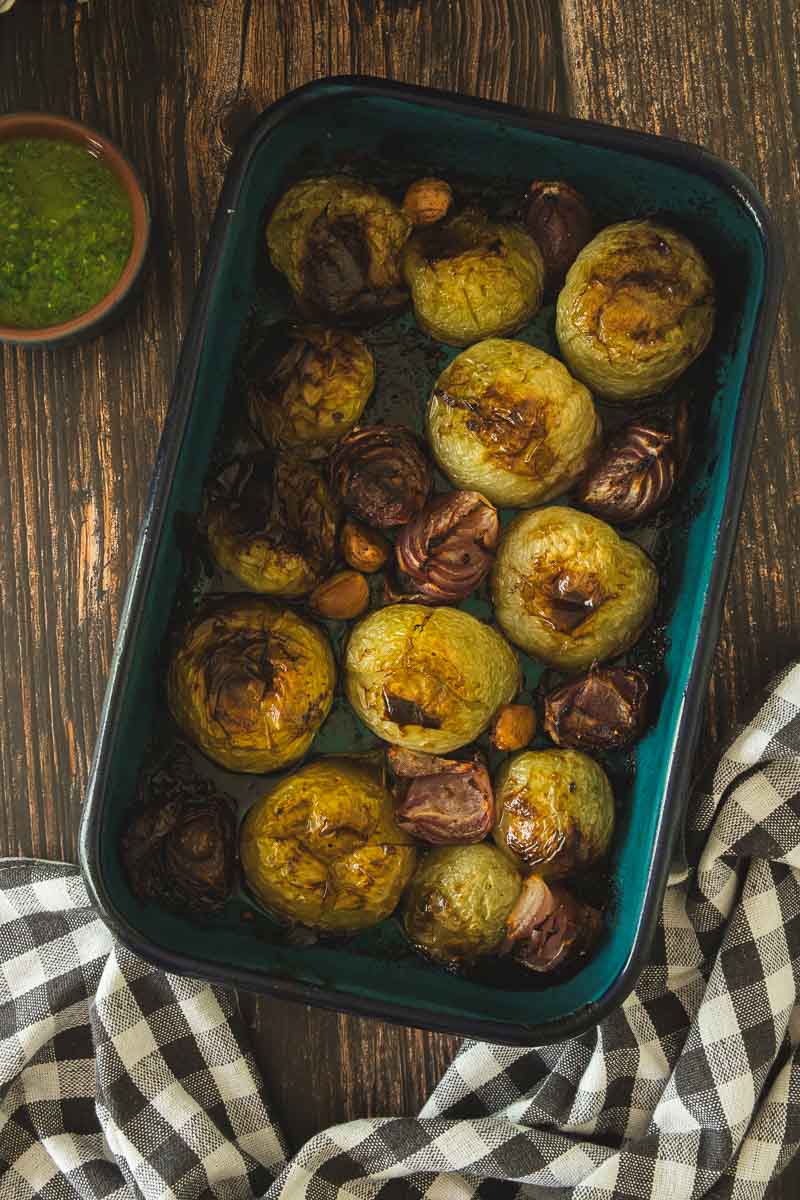 Roasted green tomatoes, onions, and garlic in a baking dish on a wooden table.