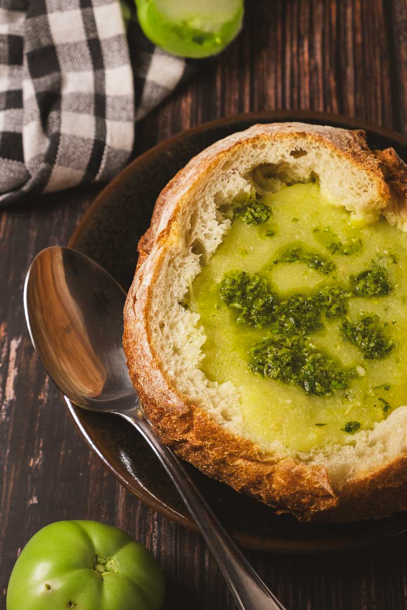 Roasted green tomato soup served in a sourdough bread bowl on a plate on a wooden table.