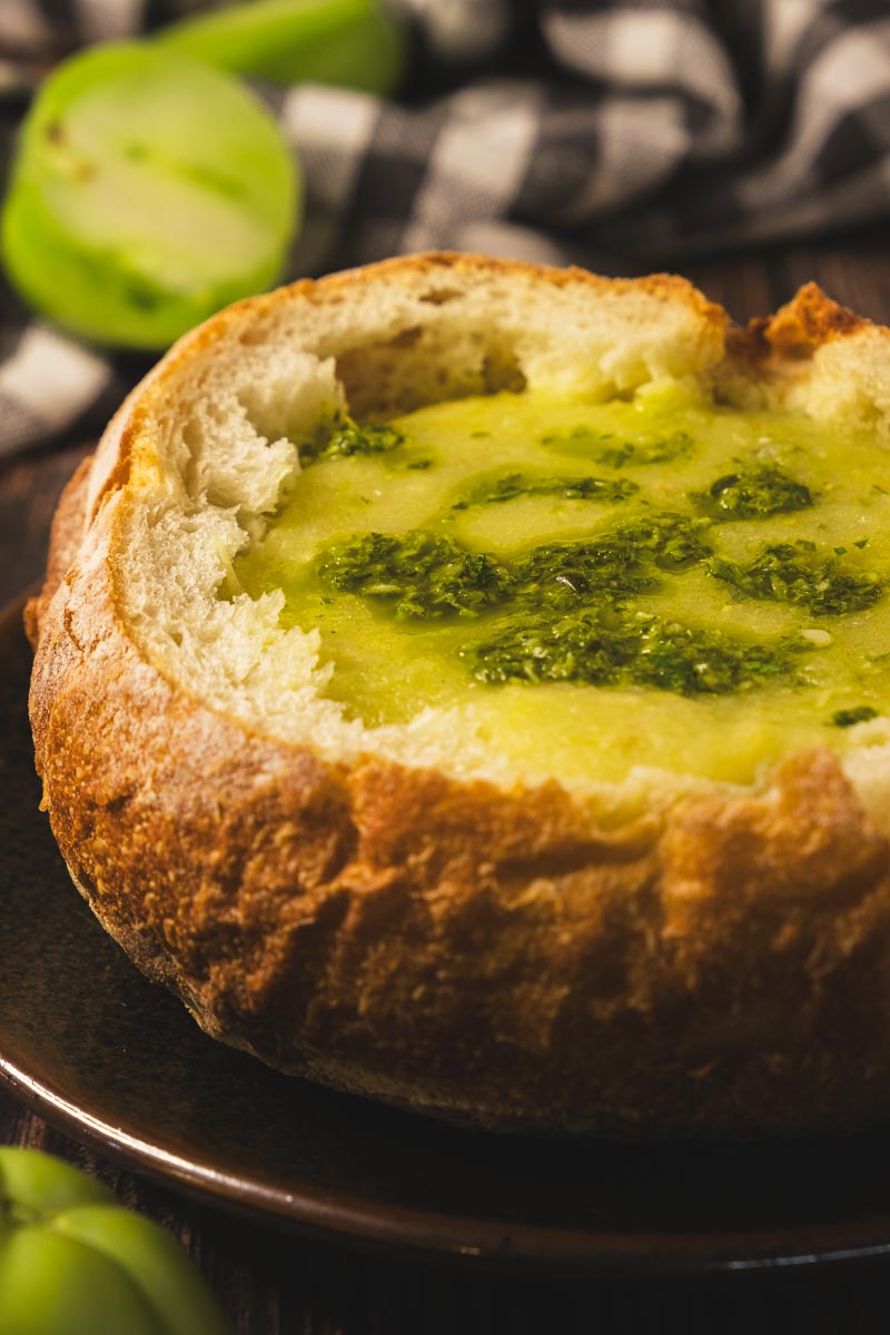 Roasted green tomato soup served in a sourdough bread bowl on a plate on a wooden table.