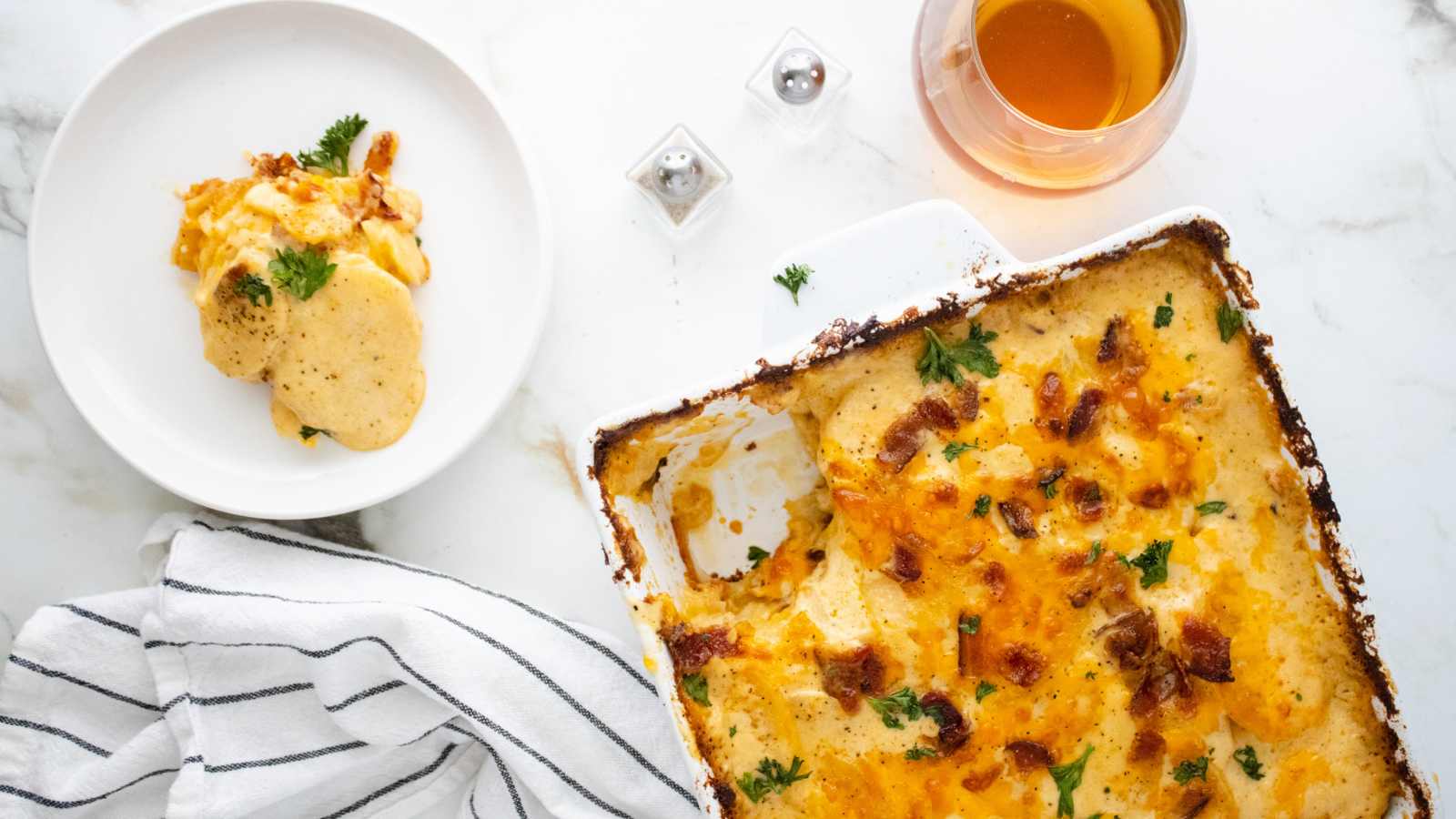 A platter full of scalloped potatoes next to a white plate with a serving.