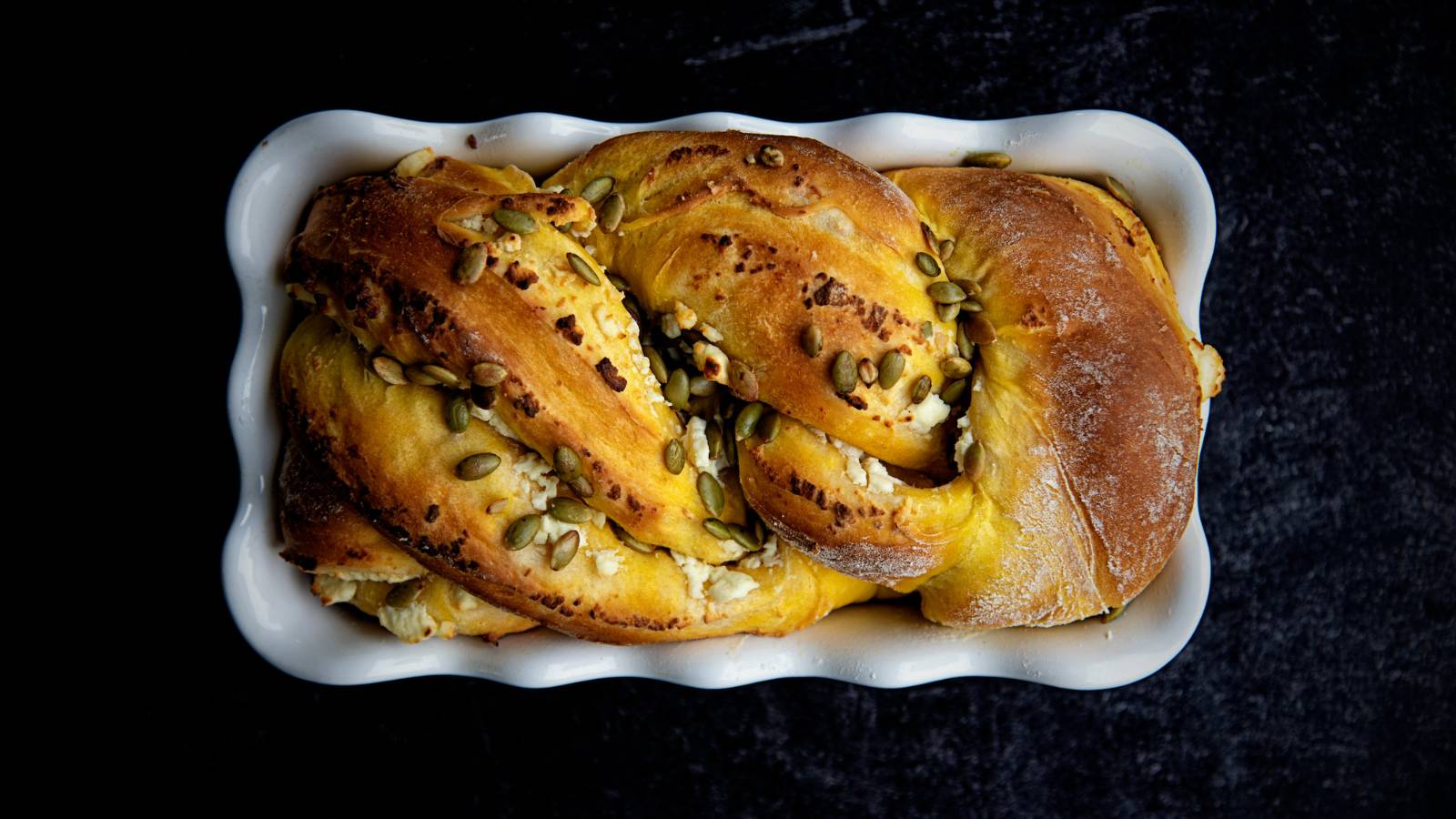 A twisted brioche loaf in a white bread pan topped with pumpkin seeds.
