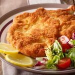 A large German schnitzel on a plate next to lemon wedges and a small garden salad.