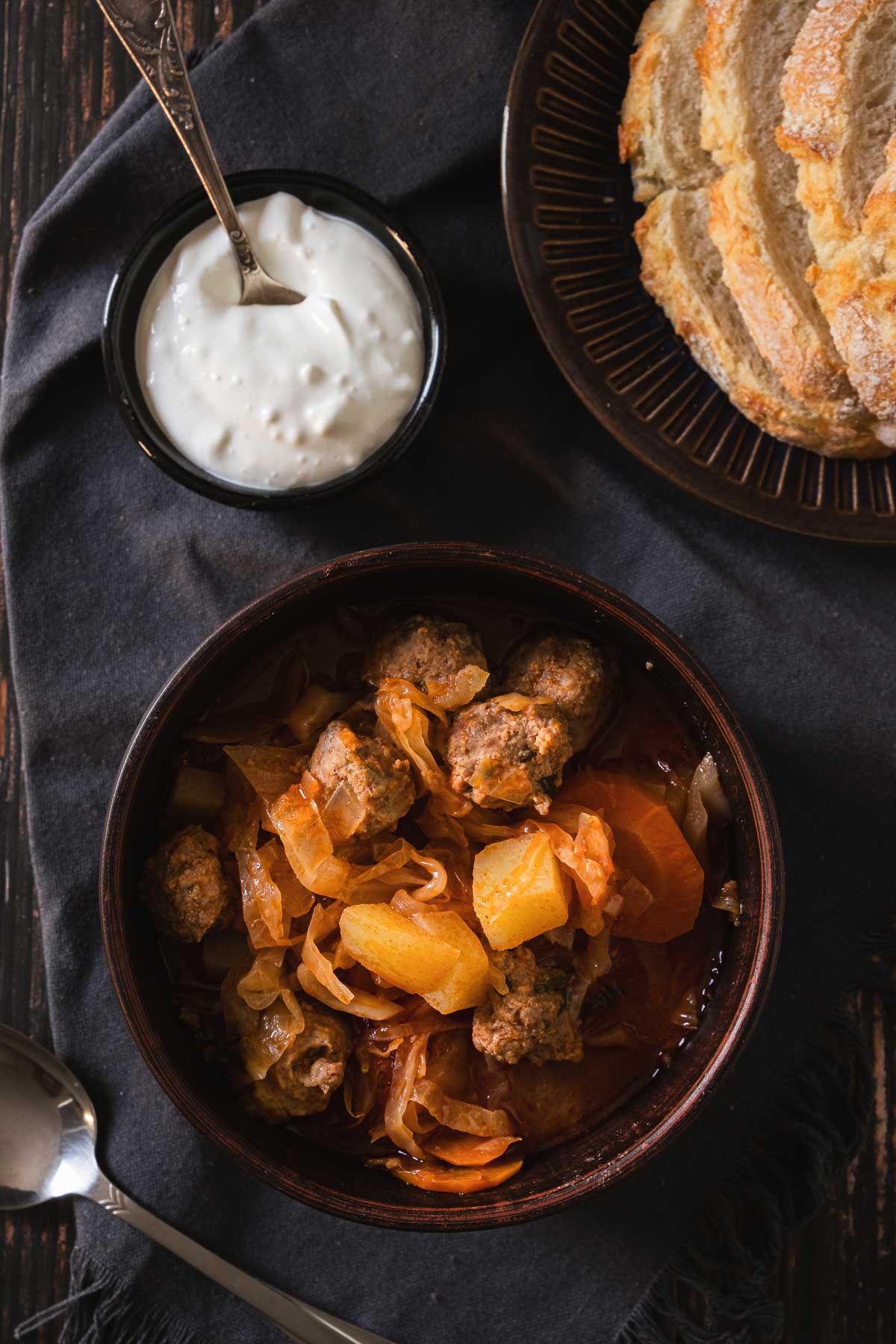 A brown bowl full of thick stew with meatballs, sauerkraut, and vegetables. Sour cream is served on the side in a separate bowl next to a basket of sourdough bread..