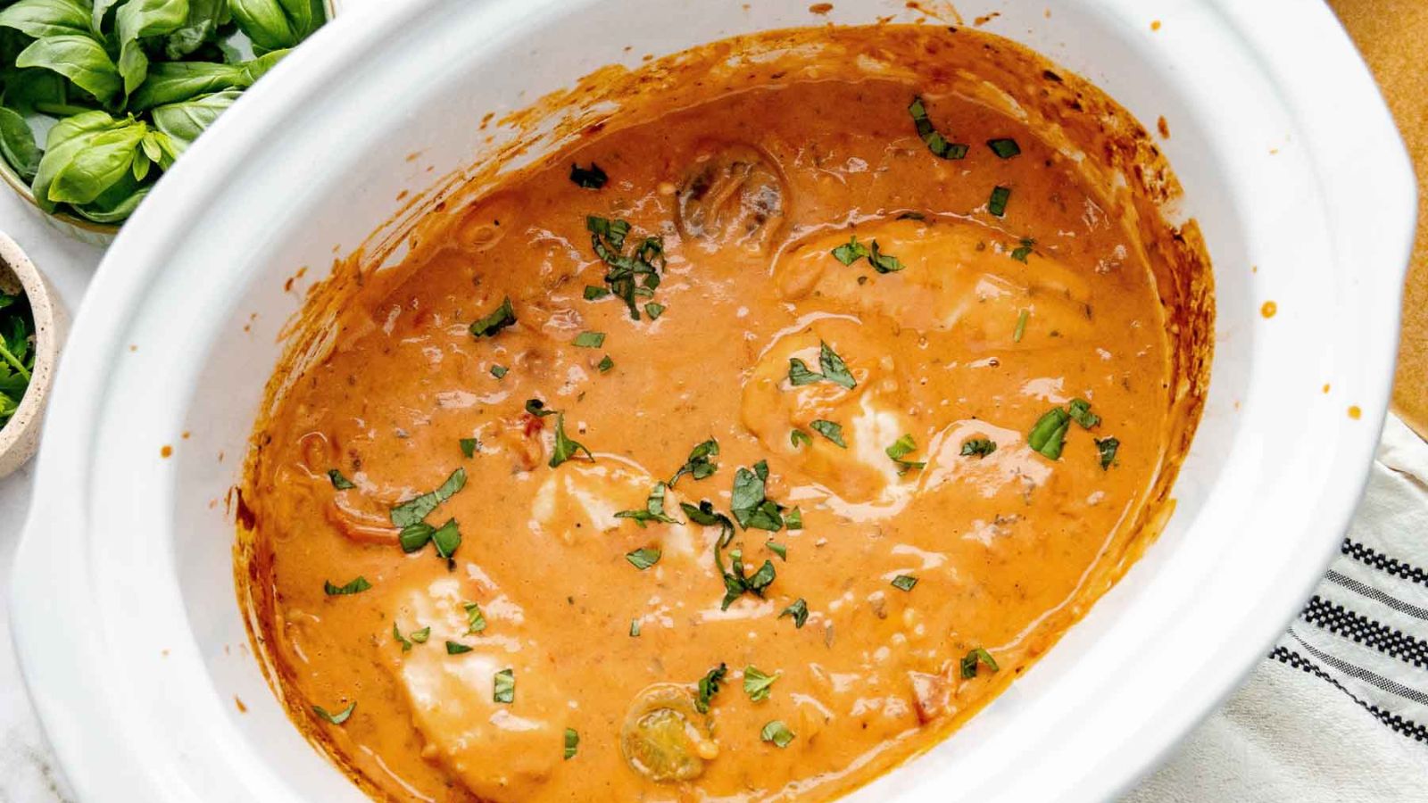 A slow cooker full of a pink, creamy tomato sauce and fresh basil smothering chicken.