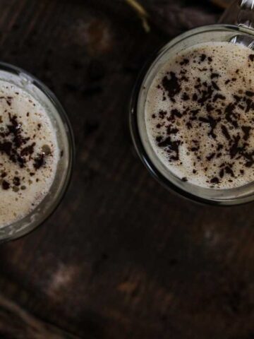 Two mugs full of frothy hot milk topped with shaved chocolate.