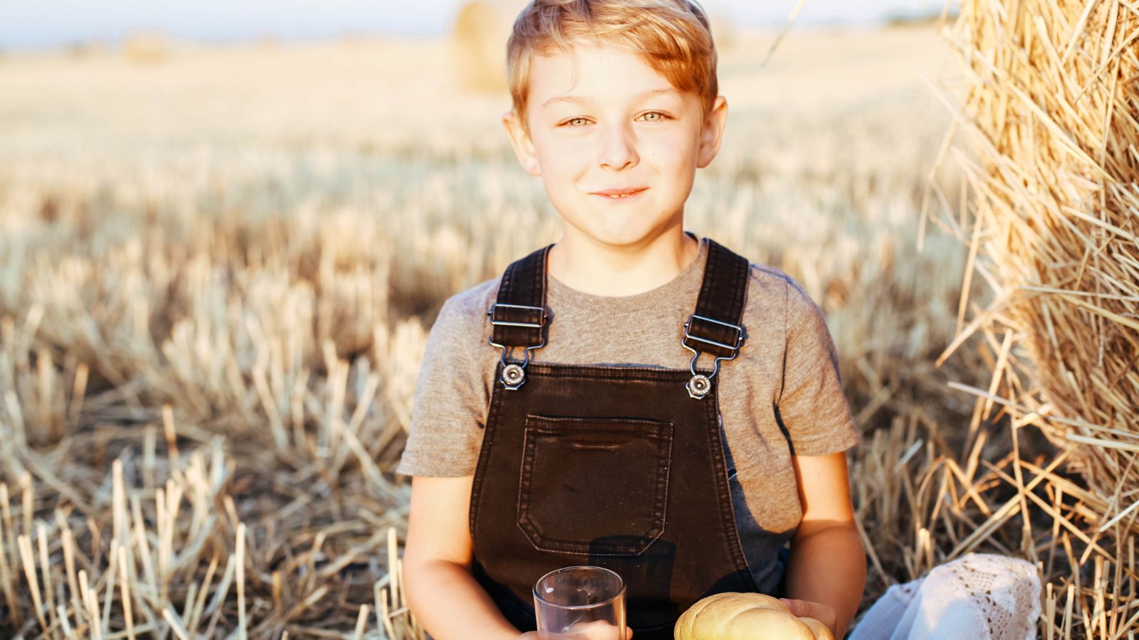 Blonde boy sitting in field next to hay.