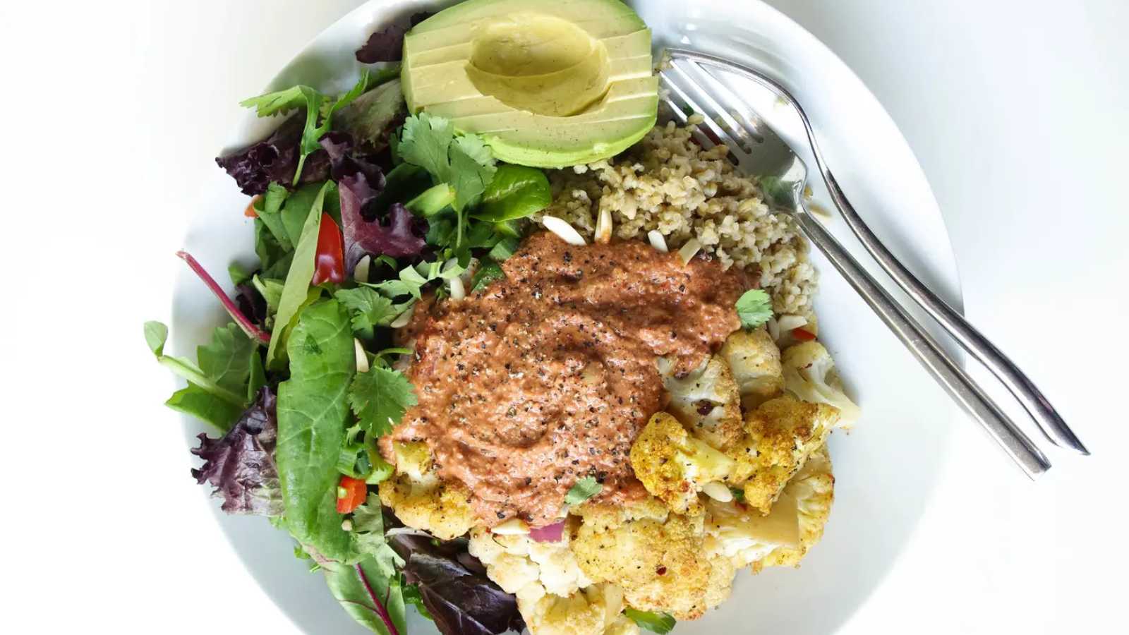 A plate of freekeh salad with sliced avocado, cilantro, green onion, and bell pepper is on one side of the bowl, while cauliflower is on the other side. In the middle, top it with a big scoop of red pepper sauce and slivered almonds.