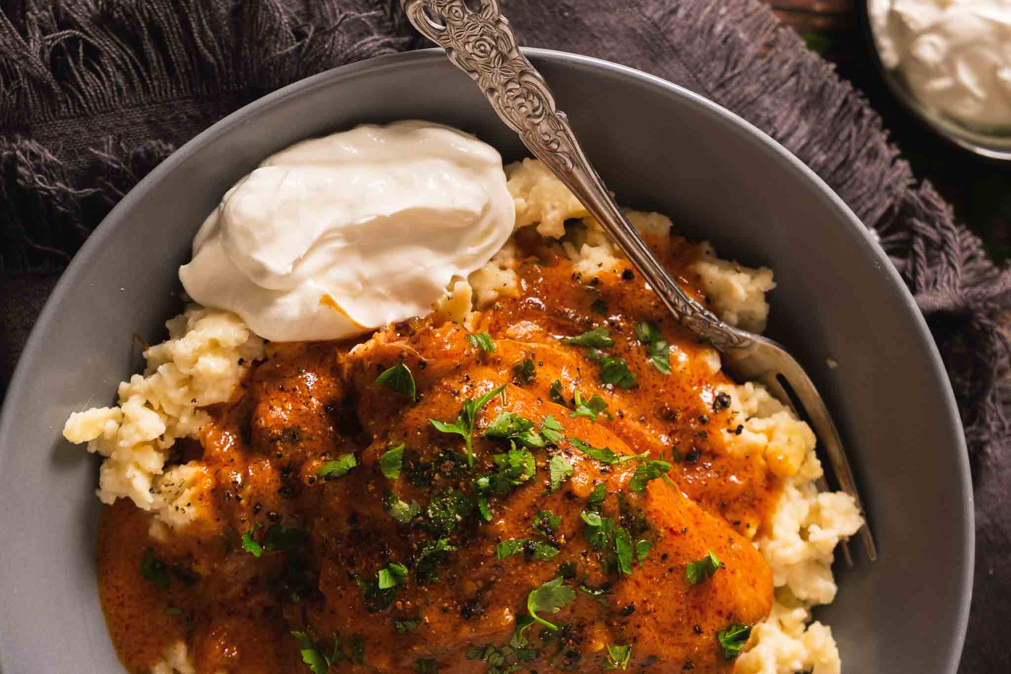 Chicken paprikash on top of spaetzle or nokedli on a plate with a dollop of sour cream on the side.