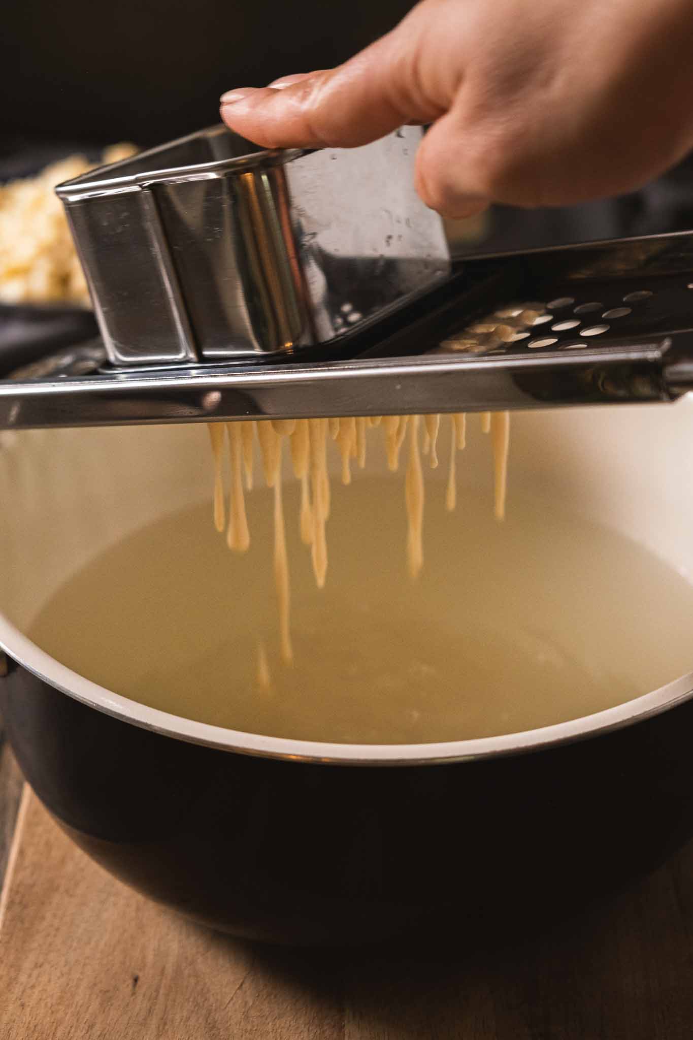 Spaetzle batter being passed through a spaetzle maker into a large pot of boiling water.